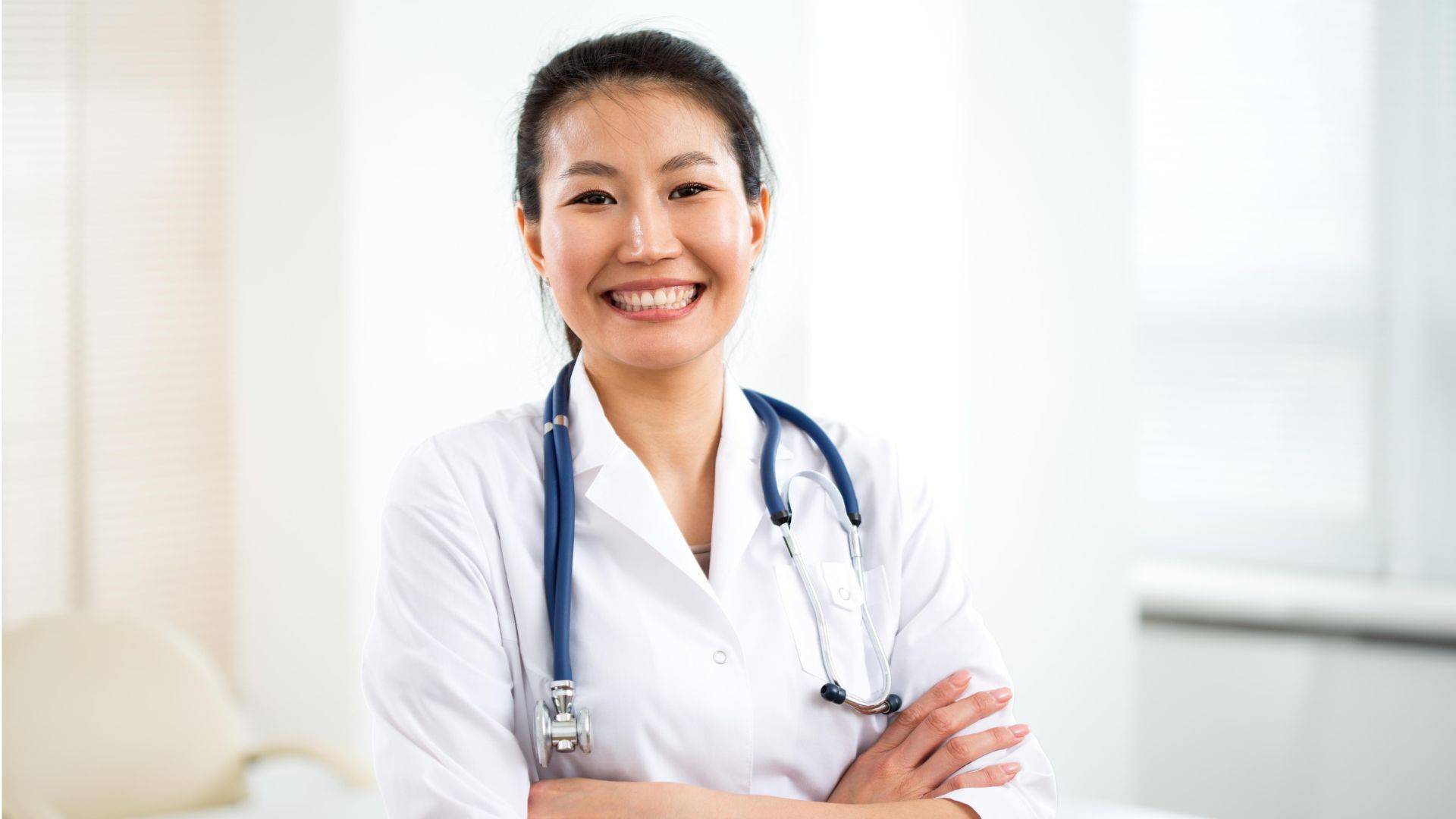 Klischeehaft gekleidete Ärztin steht lächelnd mit verschränken Armen in Behandlungszimmer