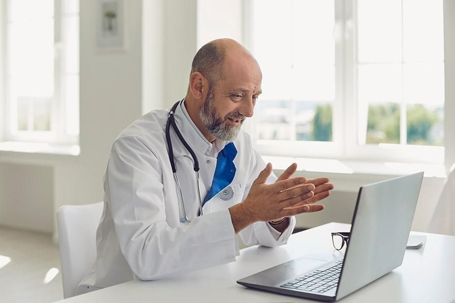 Ein Arzt sitzt in einer medizinischen Einrichtung vor einem Laptop und führt ein angeregtes Videogespräch