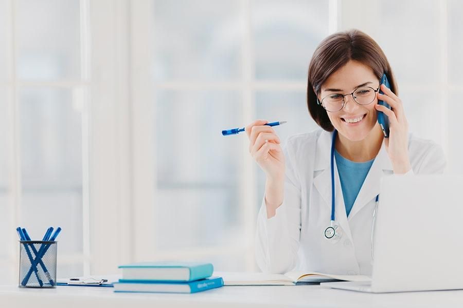 Eine Ärztin mit Stethoskop sitzt vor ihrem Laptop und führt lächelnd ein Telefonat, bei dem sie sich Notizen macht