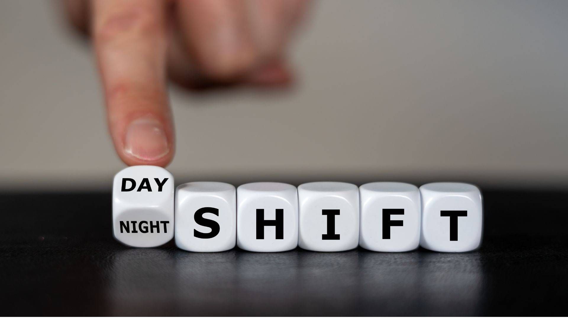 Auf einem Tisch sind Würfel platziert, welche die Wörter "Tag-" und "Nachtschicht" abbilden. Eine Hand hält den ersten Würfel in Balance.